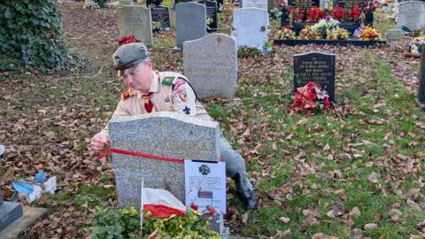 Lukasz Lakomy Lukasz Lakomy wearing a peaked cap in khaki and a light brown shirt with badges. A yellow and red tie is around his neck. He is kneeling behind a grey gravestone and tying a laminated sheet around it with a red ribbon. The grass around the grave is strewn with brown leaves. 
