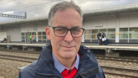 A man called Paul Gentleman who works for GWR. He is stood on a train platform and is wearing a navy blue coat over a red jumper. He is also wearing a blue collared shirt under his jumper. He has large black-rimmed glasses on.
