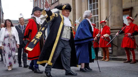 A smiling man waving his hand in the air.  He is wearing traditional mayoral dress of a long black gown trimmed with gold, a black hat and a chain around his neck.  He is surrounded by several other people including men who are dressed in red gowns. 
