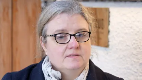 A woman with grey hair and black glasses. She is wearing a navy coat and a scarf.