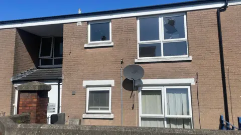 A wide view of the terraced property which was attacked in Kincraig Avenue, Belfast. The two-story building has four white framed windows. The two upstairs windows are smashed. There is a covered porch to the left and a red brick wall surrounds the property.