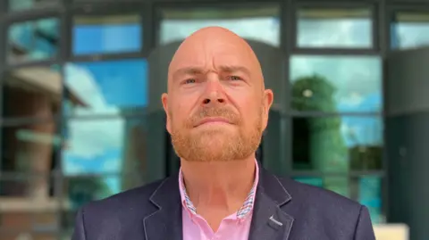 Eddie Tucker who is bald and has a light brown beard and wears a pink shirt with a black suit jacket. He is standing outside the entrance to Preston Crown Court. He looks impassively at the camera.