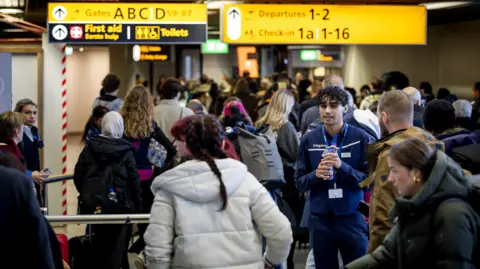 EPA/Shutterstock A queue of people at an airport, most with their backs to the camera. A man with dark hair, holding a water bottle and wearing a blue uniform, appears to be talking to a man in a brown coat. An overhead illuminated yellow and black sign to left reads "Gates ABCD, first aid, toilets", while a similar sign to the right says "Departures 1-2 Check-in 1a 1-16".