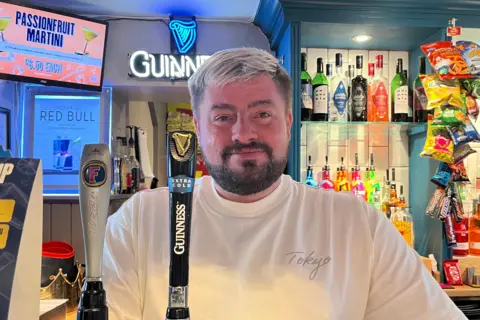 A young, bearded man stands behind a bar with bottles of alcohol in the background
