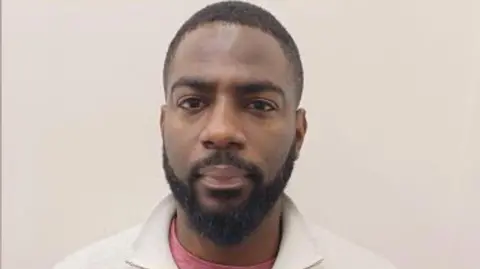 A man's mugshot. He wears a white jumper with a pink top underneath. 