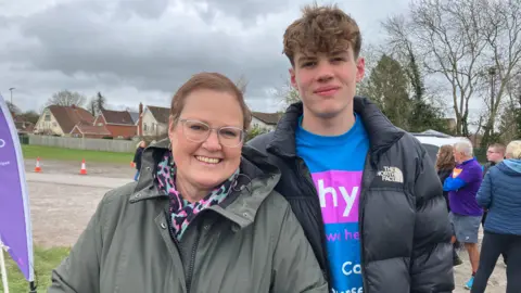 A middle-aged woman with very short red hair and clear-rimmed glasses smiles as she stands next to her 16-year-old son, who is about a foot taller than her, at a charity run. The mum is wearing a black, pink and blue leopard print scarf and a grey coat, and her son is wearing a black The North Face coat with a pink and blue charity T-shirt beneath it.
