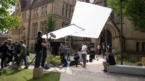 Run Away Simon Greene, played by James Nesbitt, is sitting on a wall surrounded by production equipment and crew in front of a neo-Gothic building. 