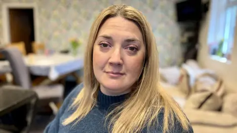 Sarra Wilson looks at the camera. She is sitting in her open plan kitchen which has been adapted for wheelchair use. She has long blonde hair and is wearing a green wool jumper.