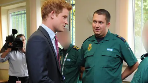 PA Media Jonathan Kirk, who has short dark hair and is wearing a green paramedic uniform, stands in front of Prince Harry, who has ginger hair and is wearing a navy blue suit and burgundy tie. 
