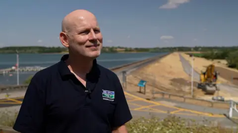 A man standing on a hillside overlooking a reservoir and embankment, with construction equipment visible in the background.