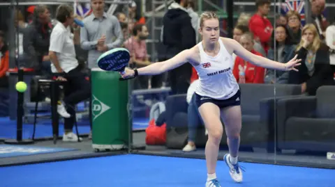 Getty Images A woman playing padel. She is wearing a white Great Britain vest and navy shorts. Spectators are watching from outside the court.