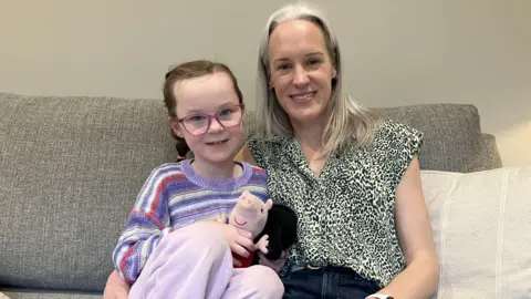BBC, Leanne Rinne A mother and daughter sat smiling on a sofa. The mother has blonde hair and has her arm cuddled around her daughter who is wearing glasses and holding soft toys in her hands. 