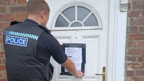 A police officer attaches a notice to a property's front door. The door is white.