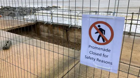 Guy Campbell/BBC Metal fencing surrounds a large, rectangular-shaped hole in a concrete promenade with a sign on the fence reading "promenade closed for safety reasons". Waves and a rock groyne can also be seen in the background. 