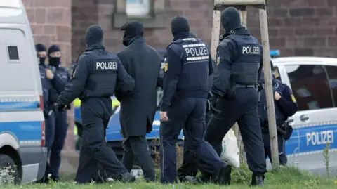 EPA Seen from behind, a group of police officers with POLIZEI written on their backs escort a man, covered in dark clothing and something covering his face, towards a police van.