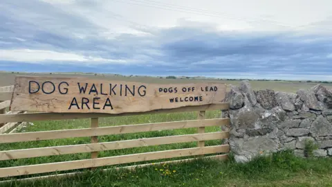 A long wooden sign on a wooden fence carries the wording: Dog walking area. Dogs off lead welcome. There is a stone wall next to it and a field beyond.