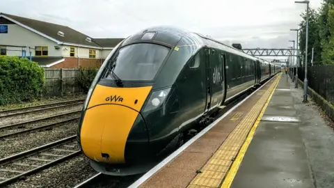 A Great Western Railway train. It has green paint and a yellow tipped nose. It is stationary by a platform. 