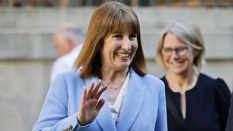 Rachel Reeves, chancellor of the UK, smiling and waving. She is wearing a light blue suit.