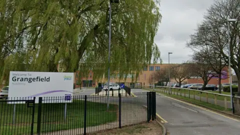 The exterior of the school from behind it's black gates. A purple and white sign on the field reads: "Welcome to Grangefield Academy". Down the driveway to the building is a car park. The orange brick building is long and three-storeys tall.