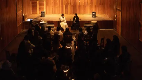 Sunah Shin A small seated audience listen to two women reading aloud as they sits under bright spotlights in the front of a gathering in a wood panelled room.
