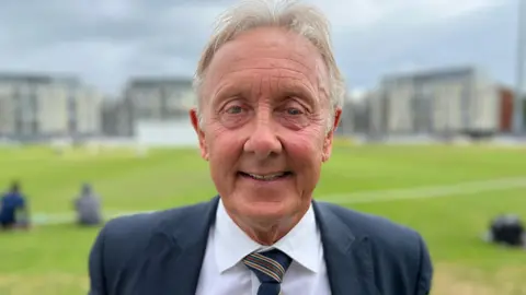 Andy Brassington. He has short grey hair and is wearing a white shirt, a tie and a grey suit jacket. He is looking at the camera and smiling