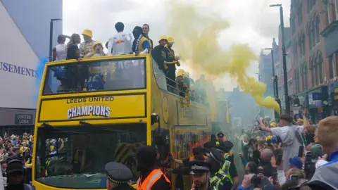 Oli Constable/BBC Leeds United bus drives along a road, with a fan waving a yellow flare among the crowds