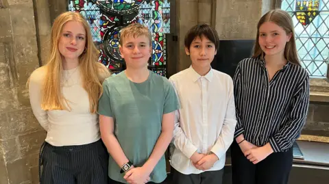 Four Canterbury Cathedral Choir members in casual clothing stood in front of stained glass window