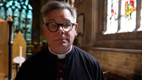 A man wearing black clerical clothing with red buttons stands inside a historic church, facing the camera. The Rev Canon Stuart Craddock is wearing round glasses and a clerical collar. Behind him are stone walls, wooden furnishings and stained‑glass windows, with daylight filtering through and illuminating the inside of St Wulfram's Church.