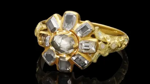 Noonans A gold ring with eight diamonds which are arranged in a floral shape. The band is engraved. The ring is against a black backdrop and is reflected in the black surface it is sat on.