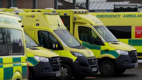 Five ambulances are lined up outside a hospital. The red 999 logo can be seen on the side of the vehicle