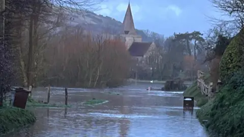 A flooded road with a large church in the background and greenery either side. 