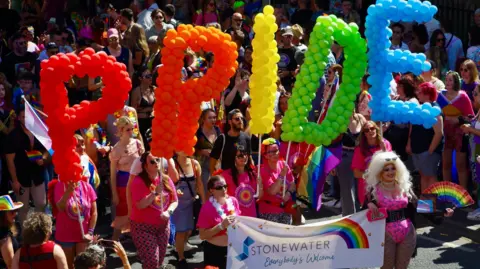 A brightly coloured parade with balloons spelling out PRIDE being carried be people in pink tops. 
