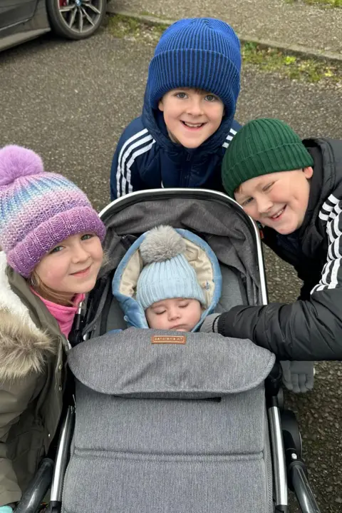 Family photo Jack is in a pushchair surrounded by his two older brothers and older sister. All are wearing bobble hats and smiling. Jack is sleeping.