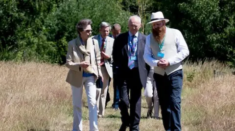 Field Studies Council A woman pinned back brown hair, wearing a pale suit and sunglasses with a handbag over her shoulder, walking in a grassy field alongside a man with a white hat and a waistcoat with a large ginger beard. Behind them there are more men in suits