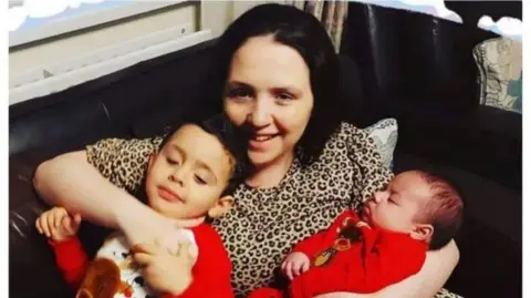 Mandy Kelly A woman and two children. The woman is wearing a leopard print t-shirt. Both children are wearing red. One child is younger than the other, and is in the woman's arms. The woman is hugging the other child with one arm. 