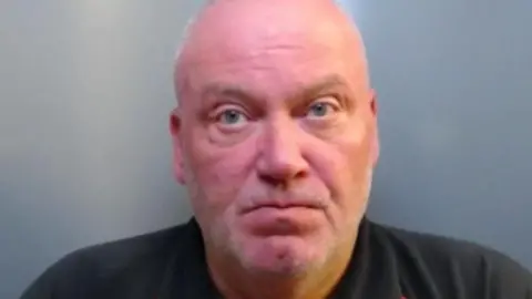 Mugshot of Ian Mcaninley. He is bald with faint grey stubble and a very red face and wearing a black polo shirt
