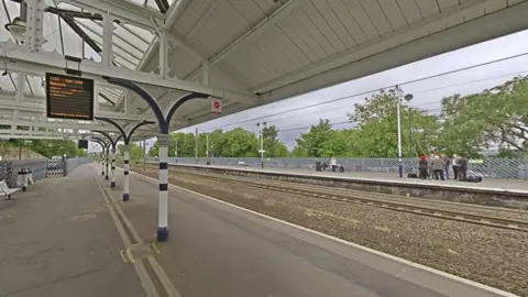 Google Northbound platform 2 at Durham station, overlooking the empty tracks. A screen is showing the next service to Newcastle. There are benches and bins. The roof of the platform is held by white and blue pillars.
A group of passengers is waiting on southbound platform 1 across the tracks. 