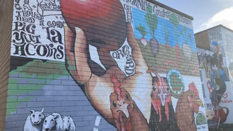 A segment of the mural that has three chicken, sheep, a hand holding an apple and some produce that is yet to be harvested