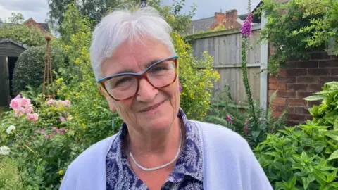 Lady smiling in a garden wearing glasses in a lilac cardigan and silver chain