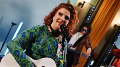 CMAT has long, red curly hair. She is singing and playing a guitar while wearing a green jumper.