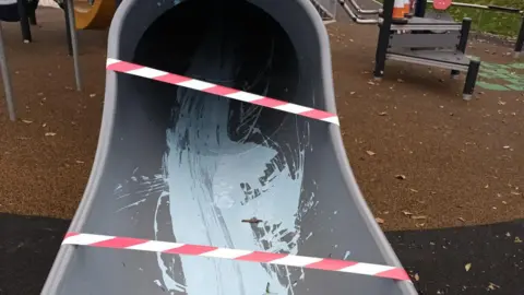 Torridge District Council The bottom of a grey tube slide with blue paint splattered across it. There is red and white tape across the bottom of it. In the background you can see brown and black tarmac as well as other play equipment