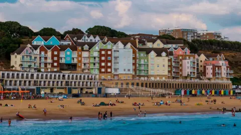 Getty Images Dozens of people lay on a sandy beach. There are colourful tall houses beyond, as well as beach huts. There are some people in the sea.