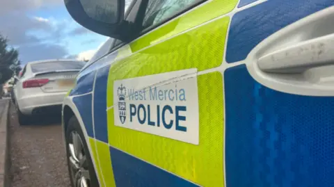 BBC The side of a white police car with yellow and blue squares on the side