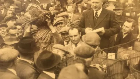 Arthur Johnson and Sons A sepia picture of a group of men in suits, caps and hats selling birds near cages. One man holds a turkey above his head