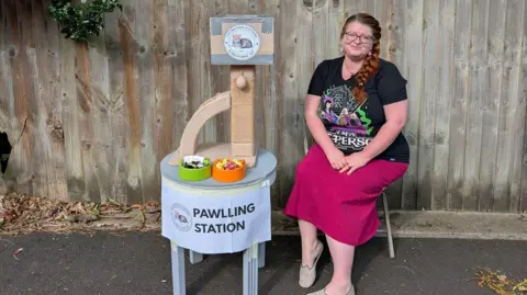 Hayley Hawkins Hayley Hawkins is sitting next to a 'Pawlling station' which consists of a table with voting slips and a cat climbing tower with a box attached to the top to put votes inside. She is wearing a black t-shirt with a colourful image on the front and a long deep pink skirt. Her red hair is tied in a very long plait which falls to the left side of her face.