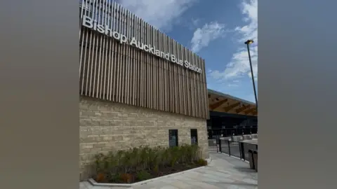 Bishop Auckland bus station is a sprawling modern building made of stone, glass and brown panels. Bishop Auckland Bus Station is written across the building in grey metal letters.