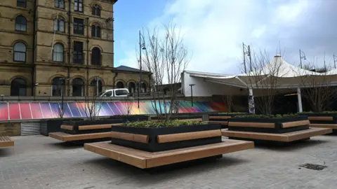 WYCA A plaza with a paved area and benches built around square planters. A white canopy is in the background and a large building to the left.