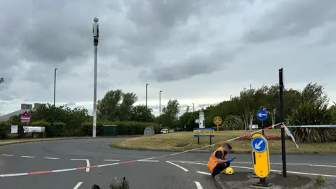 Burned white telephone mast next to roundabout with grass on it. Surrounding it are police tape and a man with a high-vis orange jacket checking something on the ground