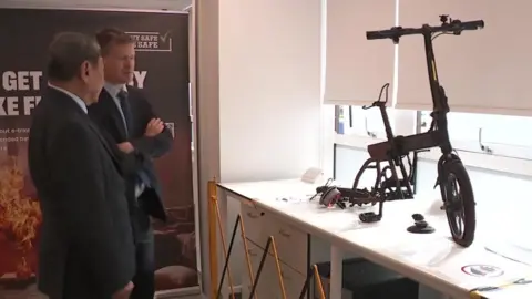 Product Safety Minister Justin Madders (right) stands and chats with another man while looking at a burnt out e-bike on display in an office