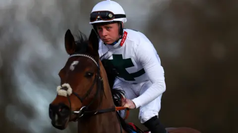 Getty Images Levi Williams sitting on a brown horse, wearing an all-white outfit, with a white helmet and black glasses. He is holding on to the reins of the horse.